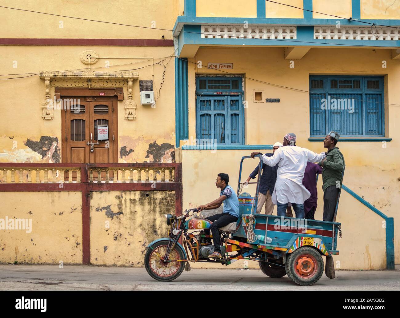 A three wheeler tempo rickshaw rides through a street past old houses with quaint blue and brown wooden doors in the island of Diu. Stock Photo