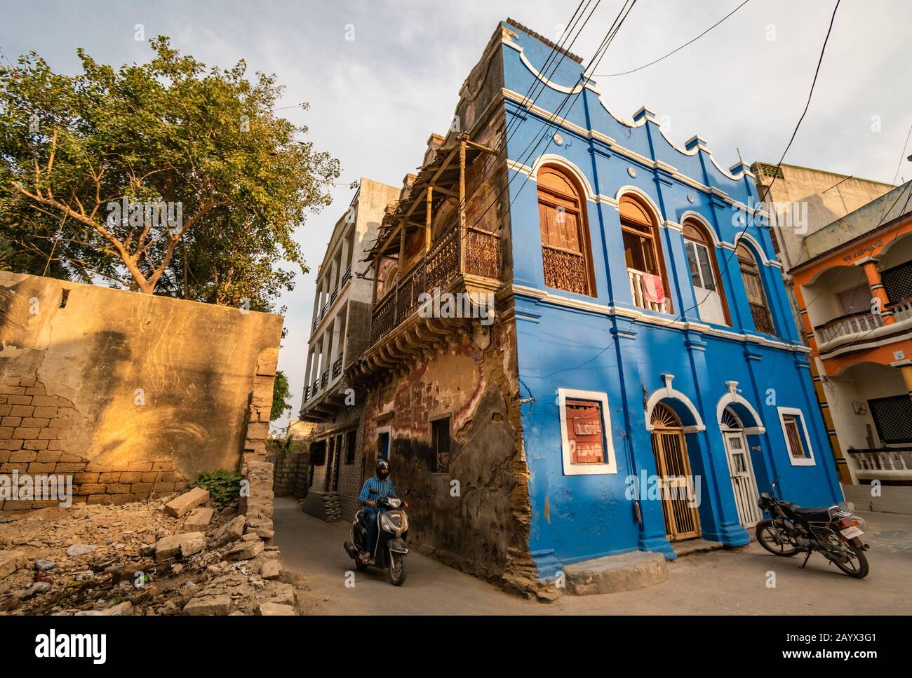 A man bikes through the narrow lanes of the old town of Diu. An old, colonial era house with blue walls and an ornate balcony looms over the street. Stock Photo