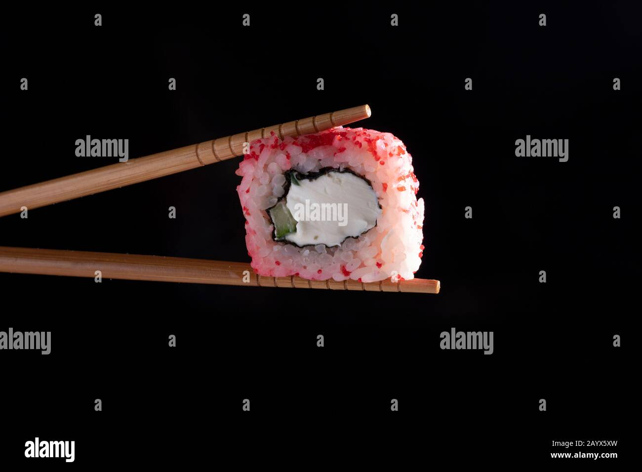 Chopsticks holding seaweed pink roll filled with avocado and fish over black background Stock Photo