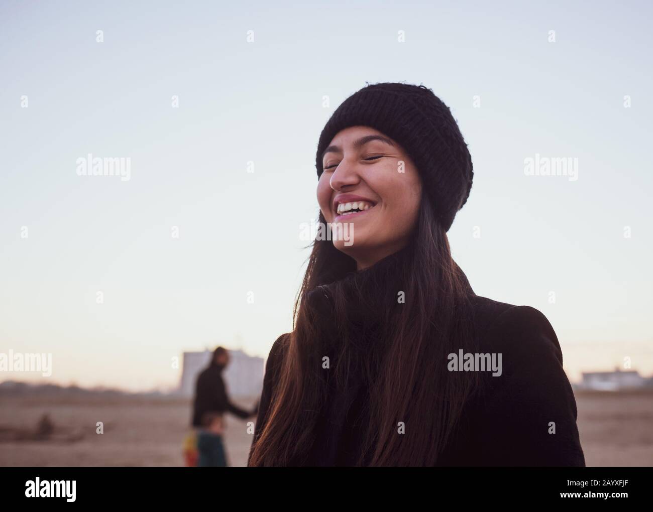Young woman in winter clothing with wool hat smiling with closed eyes, emotion of authentic joy. Location on the beach with twilight light. Stock Photo