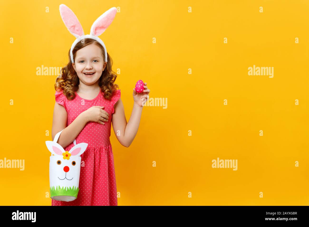 A beautiful cute little girl in the ears of a bunny holds a bag for sweets and an avshalny egg. Portrait of a child on a yellow background. Copy space Stock Photo