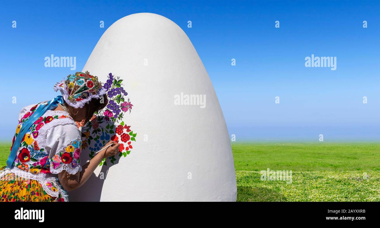A giant white Easter Egg is being decorated with folklore floral patterns by a woman wearing traditional embroidered folk costume. Stock Photo