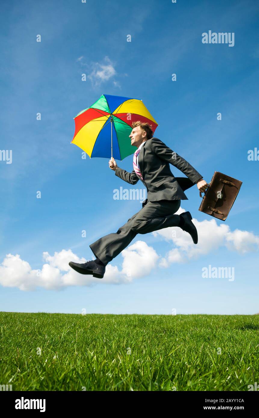 Office worker with colorful umbrella and briefcase jumping outdoors across field of green grass Stock Photo