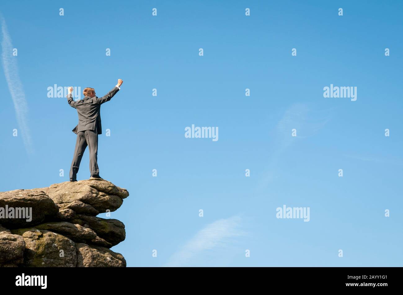Businessman standing on the top of a distant rocky mountain peak celebrating in blue sky copy space Stock Photo
