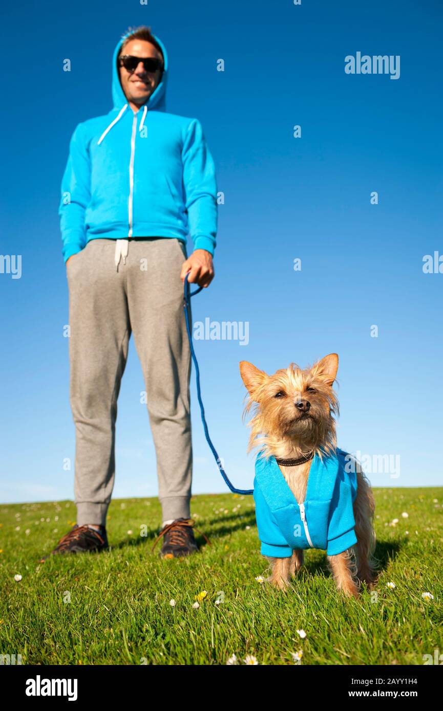 Small fluffy dog standing with his best friend owner in matching blue hoodies outdoors on bright green field Stock Photo