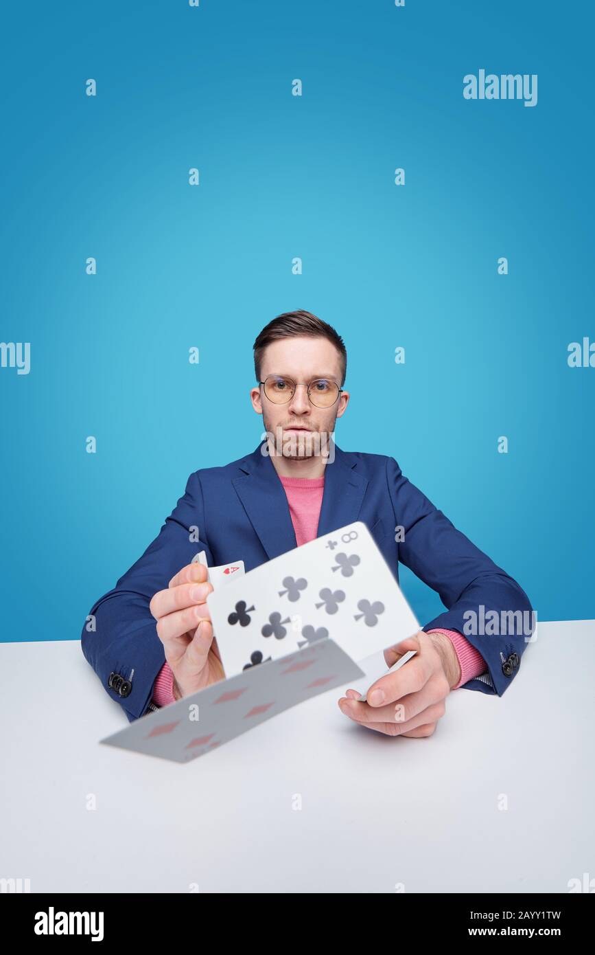 Portrait of serious intelligent young bearded card player in glasses sitting at table and throwing cards to camera Stock Photo