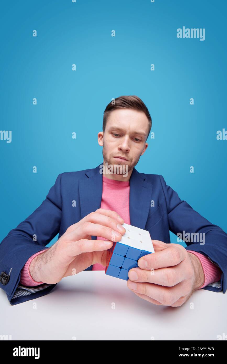 Serious young man in jacket sitting at table and solving cube puzzle while developing logic Stock Photo