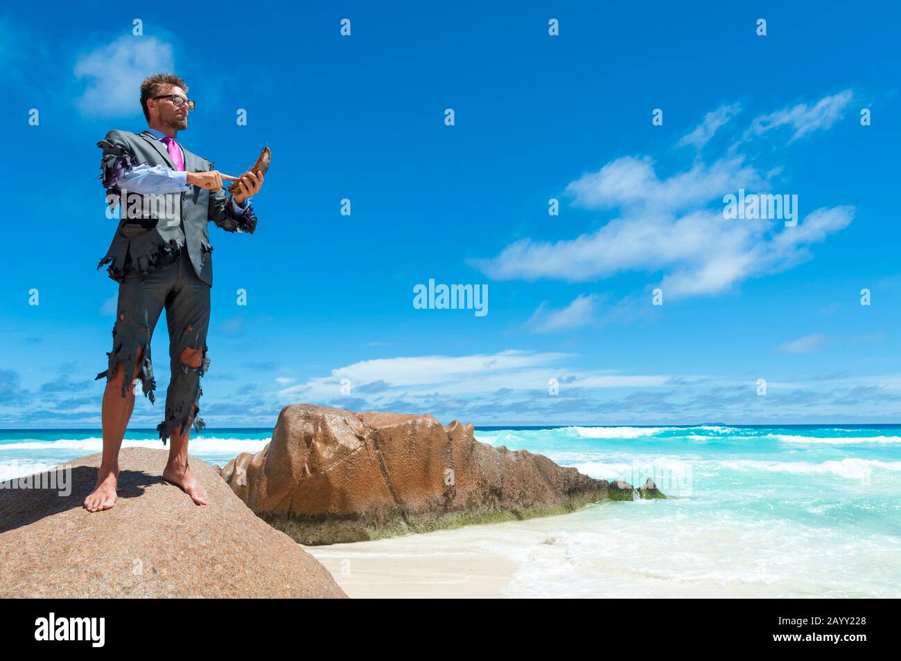 Survivor businessman in tattered suit standing on rough rock using coconut husk mobile phone on tropical beach Stock Photo