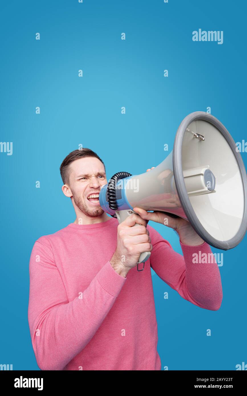Young man with expressive face speaking announcement into megaphone against blue background Stock Photo