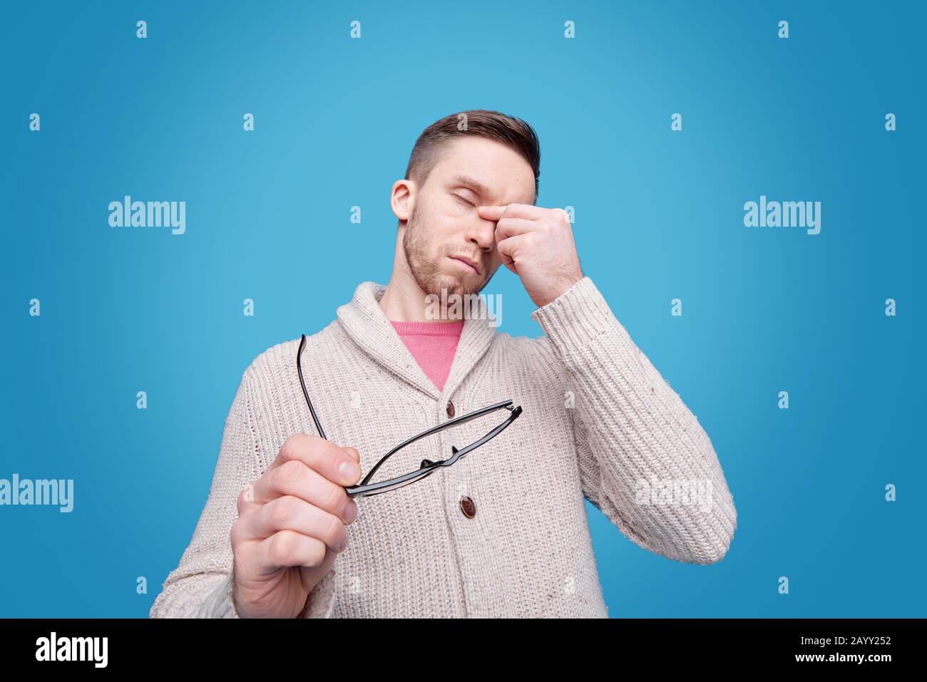 Tired young bearded man in cardigan rubbing bridge of nose and holding glasses against blue background Stock Photo