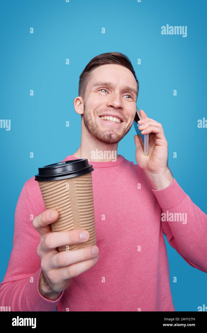Smiling handsome young man with stubble drinking coffee from takeout cup and chatting by cellphone Stock Photo