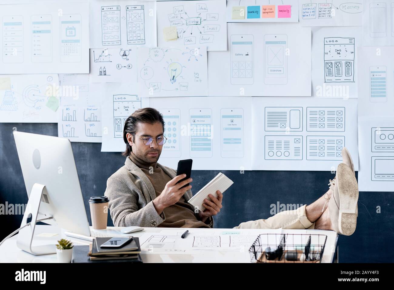 Stylish Caucasian man sitting relaxed at his office desk with feet on it holding smartphone at tablet PC Stock Photo