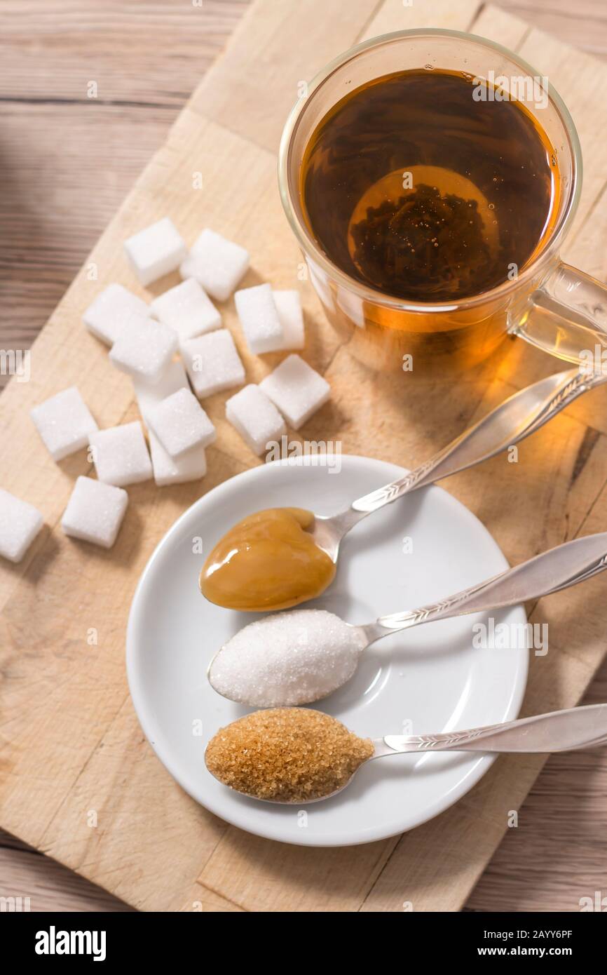 Lump sugar, cane sugar, loose white sugar and buckwheat honey on a teaspoon lie next to a glass with brewed herbal infusion. Stock Photo