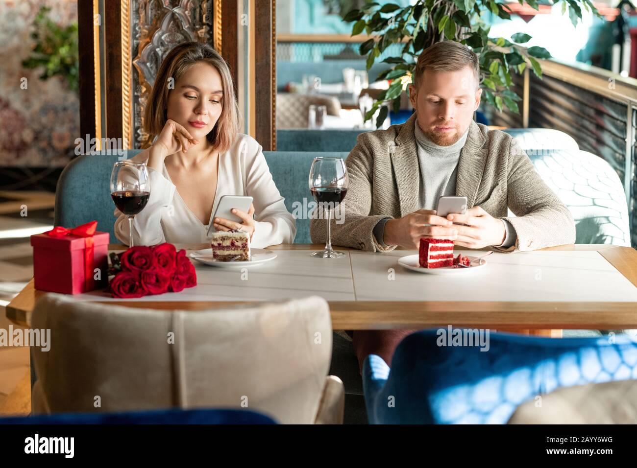 Young adult Caucasian man and woman sitting together in modern restaurant surfing Internet on their smartphones instead of talking to each other Stock Photo