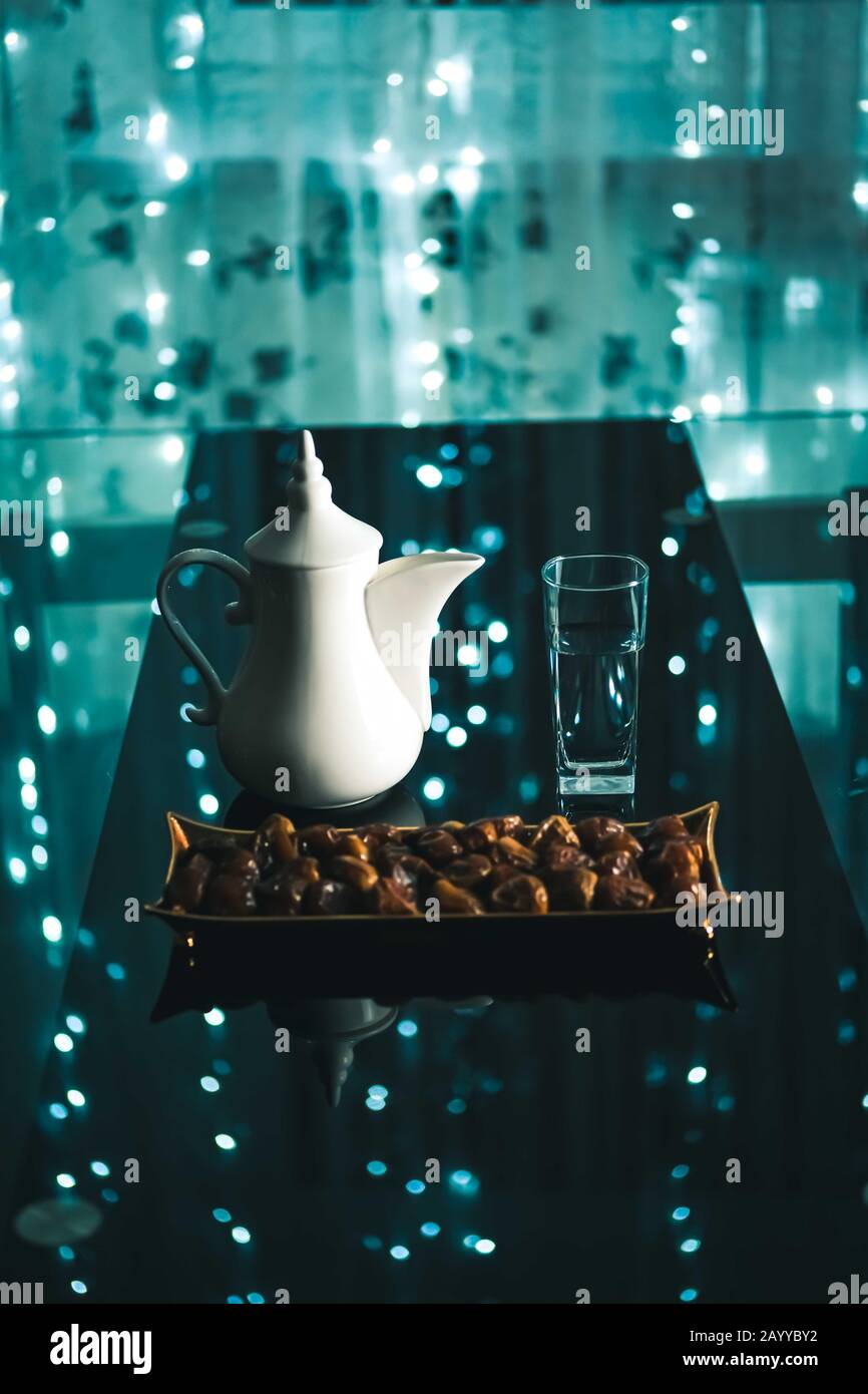 Beautiful bowl in Arabic style full of dry dates fruits, white tea pot and a glass of water, Ramadan concept, on dark blue background and fairy lights.Vertical. Stock Photo