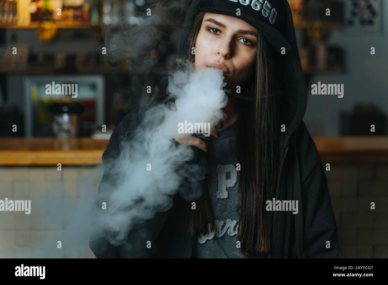 Young pretty woman in cap smoke an electronic cigarette at the vape shop. Hip-hop style. Closeup. Stock Photo