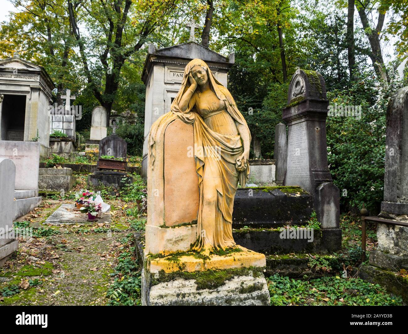 Old tombstone covered with musk with a statue of a young woman crying over a plaque in a path under the trees of monumental cementery Stock Photo