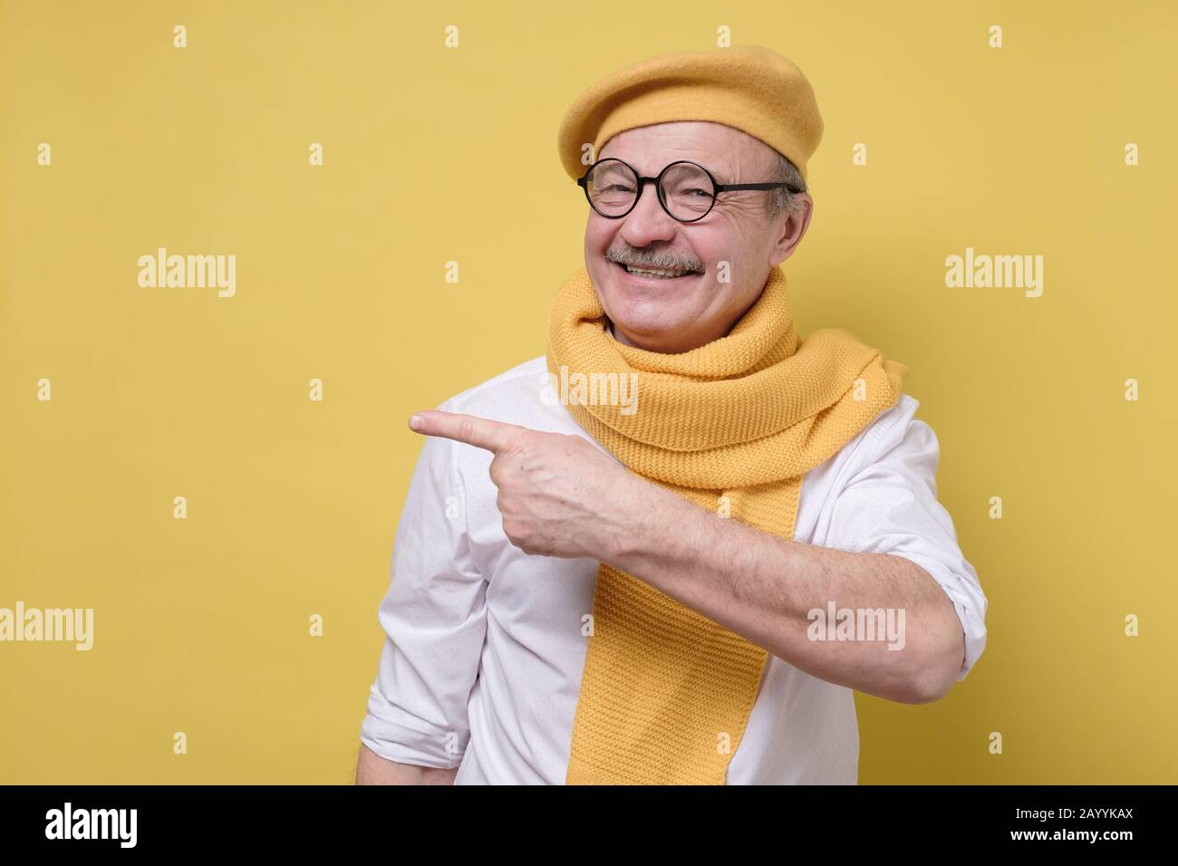 Confident senior smiling man in yellow beret and scarf point away. Serious mature male gesturing with his finger aside on yellow background. Stock Photo