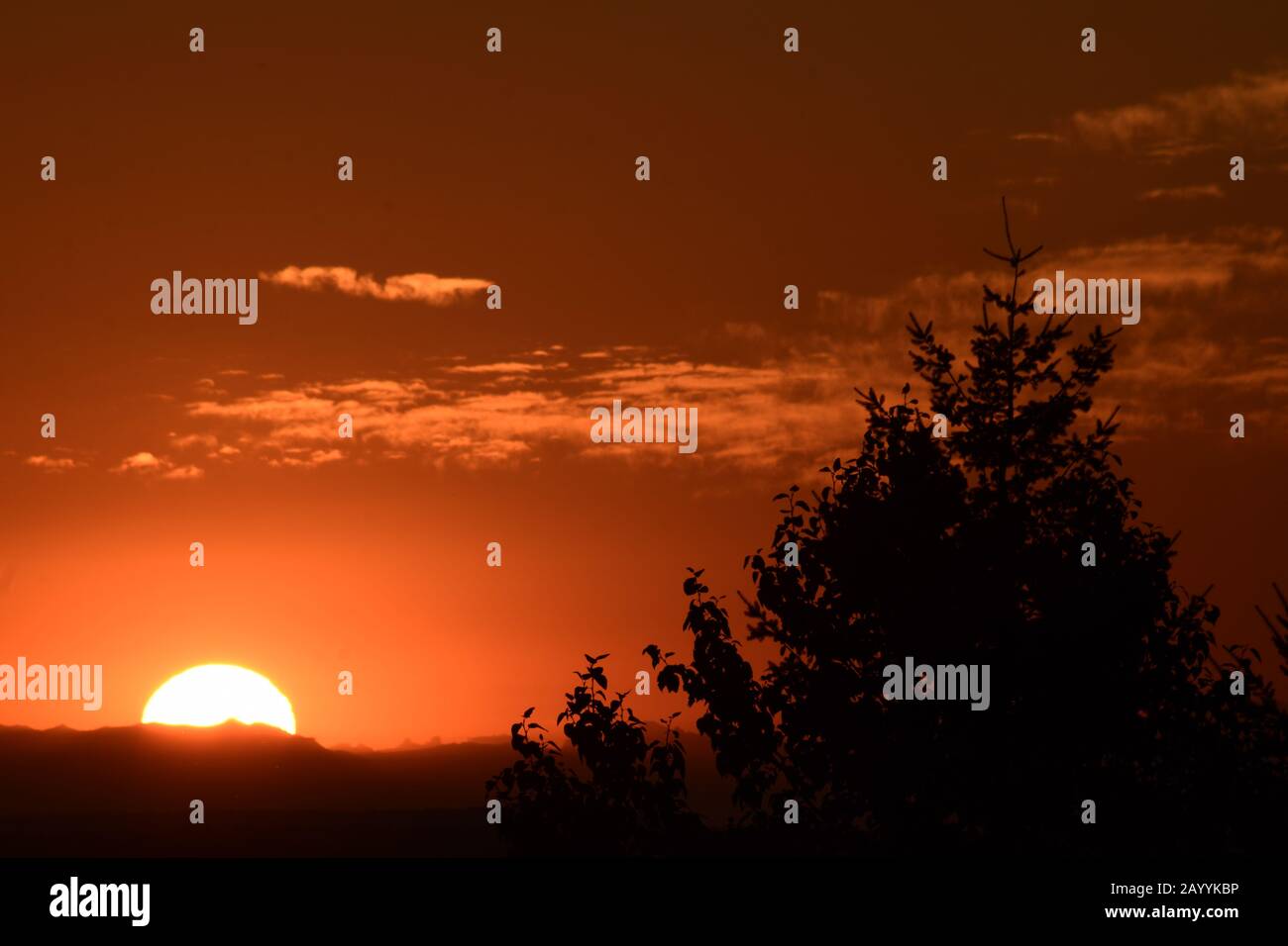 Bright Sun Rising Over Dramatically Silhouetted Mountain Ridge Against Orange Cloudy Sky Stock Photo