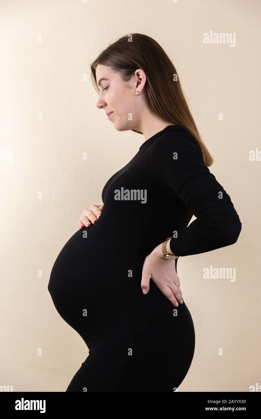Side view of an attractive pregnant woman caressing her belly in black dress  while standing against yellow vertical background Stock Photo - Alamy