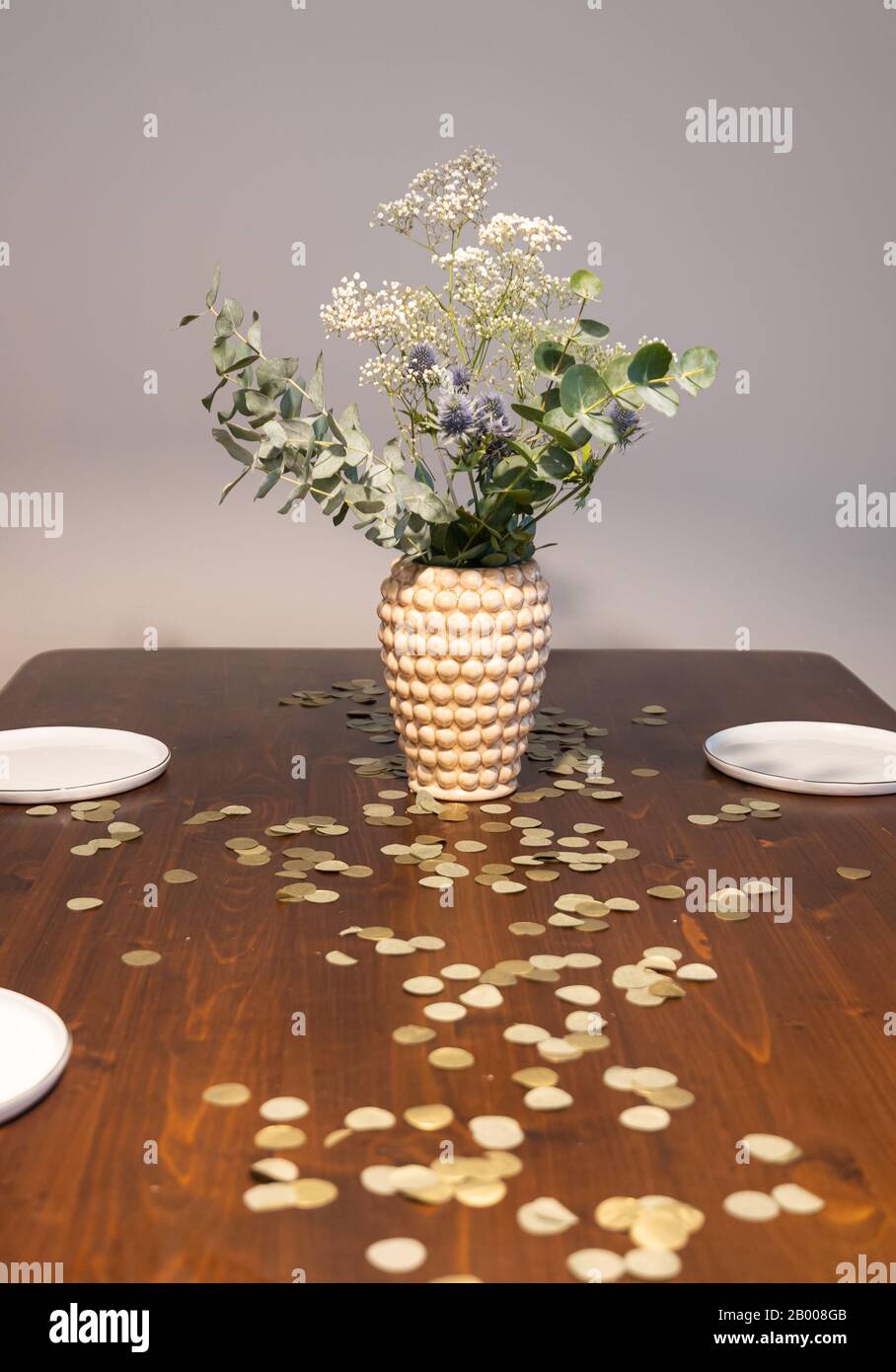 Vase with a bouquet of white and blue flowers on a wooden table before a grey background. golden confetti lying on the table. Stock Photo