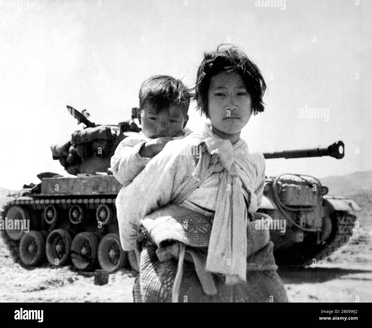 Korea: With her brother on her back a war-weary Korean girl  trudges by a stalled M-46 tank at Haengju, Korean War (1950-1953), 9 June 1951.  The Korean War (25 June 1950 - armistice signed 27 July 1953) was a military conflict between the Republic of Korea, supported by the United Nations, and North Korea, supported by the People's Republic of China (PRC), with military material aid from the Soviet Union. The war was a result of the physical division of Korea by an agreement of the victorious Allies at the conclusion of the Pacific War at the end of World War II. Stock Photo
