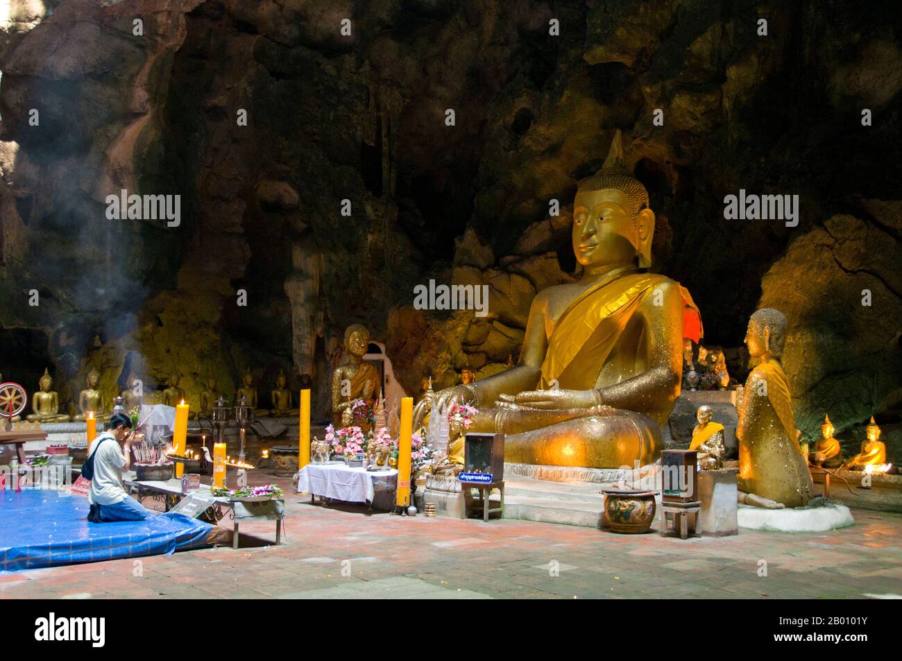 Thailand: Worshipper in front of a large seated Buddha inside Tham Khao Luang, Phetchaburi.  Tham Khao Luang is a large cave consisting of three linked chambers and is distinguished by many hanging stalactites and Buddha images, including a phra non or reclining Buddha. The main bronze image was cast on the orders of King Chulalongkorn (Rama V) and dedicated to his illustrious predecessors, Kings Rama III and Rama IV. There is a natural opening in the ceiling of the second, main chamber, through which daylight streams illuminating the images within. Stock Photo