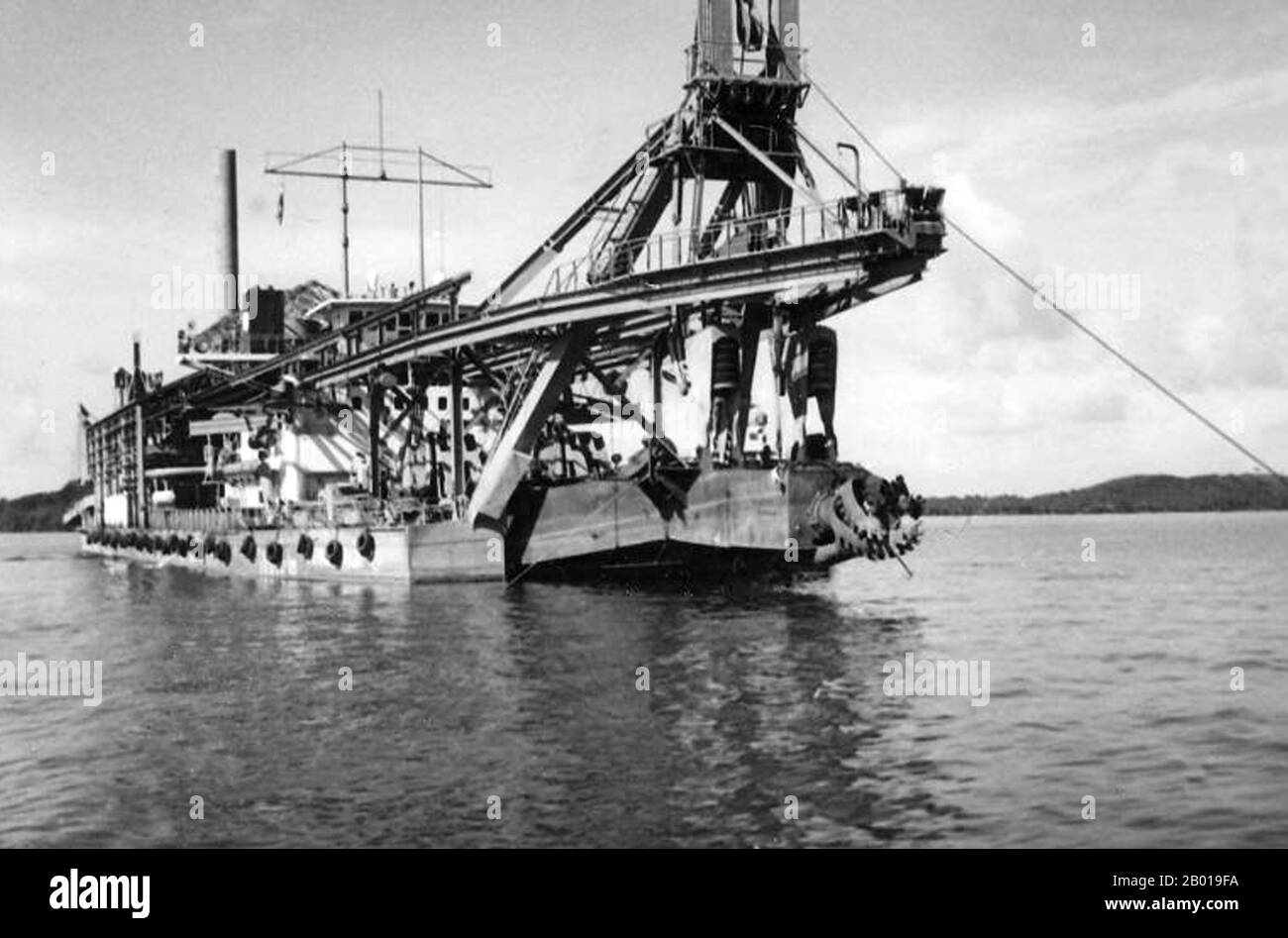 Thailand: A tin dredger anchored off Phuket in the Andaman Sea c. 1915.  The introduction of the first tin dredger in 1907 allowed the tin mining industry on Phuket to expand into a vast new area that had previously been untouched. Several types of dredges were used locally. Hydraulic dredges sucked the ocean floor for the alluvial deposits of tin through a pipe, separated the tin and discharged the spoil on the shore through a floating pipeline. Elevator dredges employed an endless chain of small buckets to scrape the ocean floor and separate the tin ore from the rest of the spoil. Stock Photo