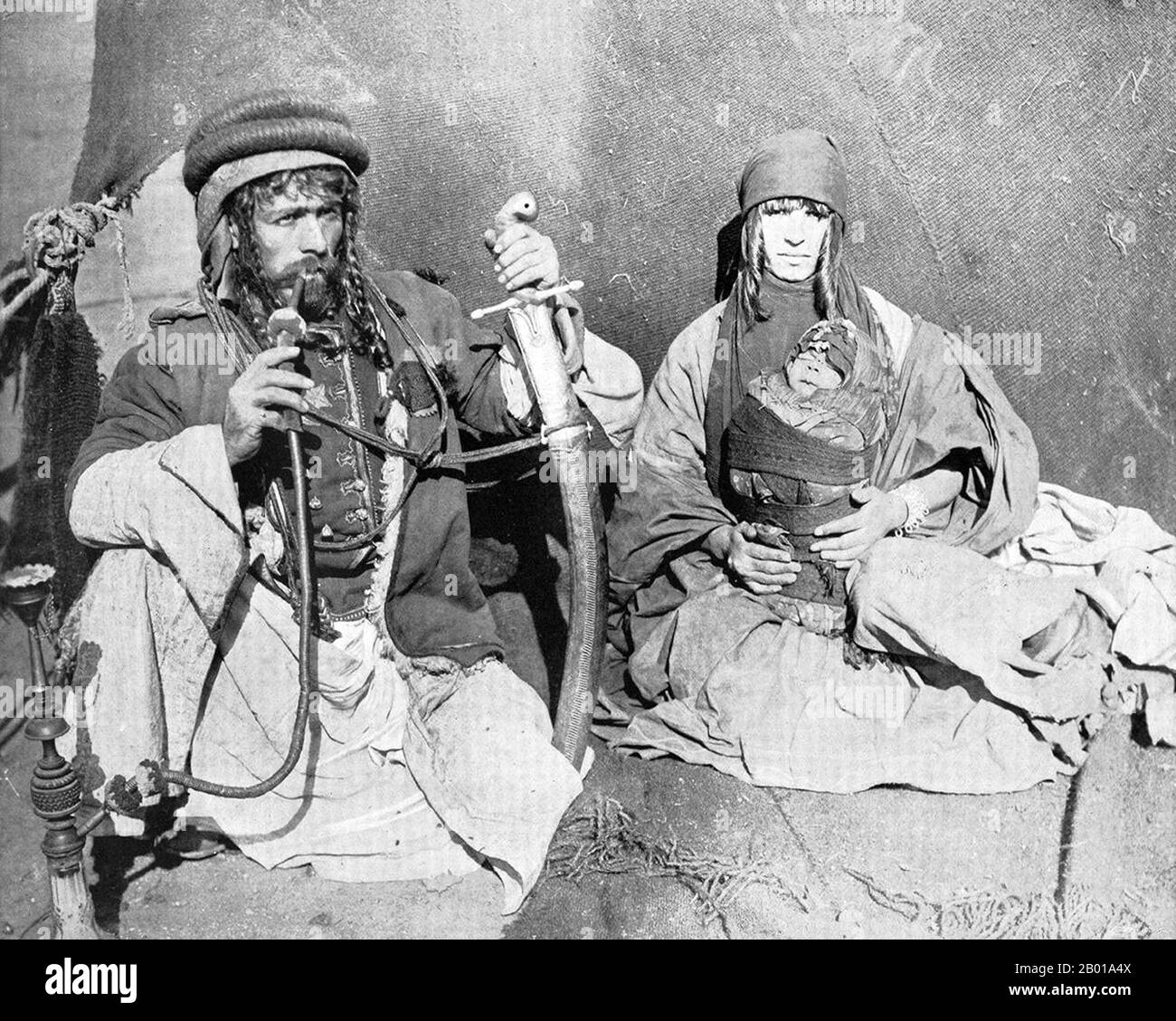 Syria: A Syrian Bedouin with his wife and tightly-swaddled child, 1893.  The Bedouin (from the Arabic badawi, pl. badawiyyūn) are a part of the predominantly desert-dwelling Arab ethnic group. Specifically the term refers only to the 'camel-raising' tribes, but due to economic changes many are now settled or raising sheep. Also due to linguistic and cultural changes the term is now often applied in many ways either to Arabs in general or to desert dwellers or nomads. In Syria, the Bedouin live predominantly in the desert east and south of the country. Stock Photo