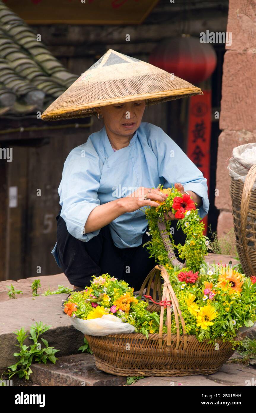 China: Tujia woman selling flower tiaras at the North Gate Tower, Fenghuang, Hunan Province.  Fenghuang is Chinese for Phoenix and refers to the mythical sacred firebird that can be found in the mythologies of the Persians, Greeks, Romans, Egyptians, Chinese, and (according to Sanchuniathon) the Phoenicians.  Legend suggests that two phoenixes on discovering the town hovered overhead for some considerable time before reluctantly flying away.  Fenghuang town is a well-preserved ancient town supposedly dating back to 248 BC. It is home to the Miao and Tujia minorities. Stock Photo