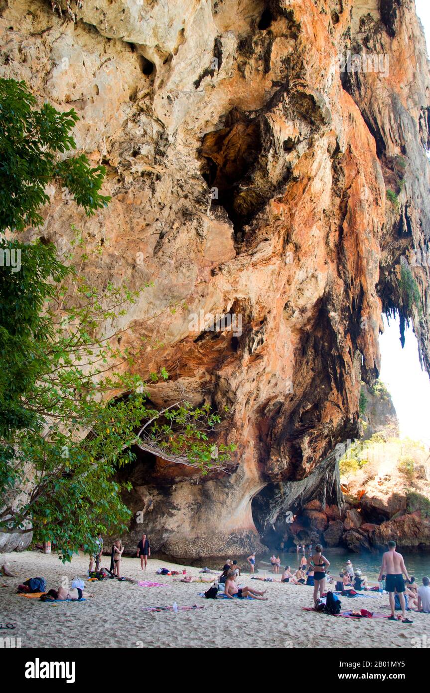 Thailand: Enjoying the shade of the cliff next to the Phra Nang cave, Tham Phra Nang, Krabi Coast.  Tham Phra Nang or the ‘Cave of the Revered Lady’ is located near the southern tip of the narrow peninsula that divides the beaches of Rai Leh East and Rai Leh West.  Dedicated to the memory of an eponymous Indian princess said to have been drowned offshore centuries ago, a fertility cult has developed around the cave and a small shrine within. The cave itself is packed with red-tipped phalluses placed here by fishermen in the hope of good catches. Stock Photo
