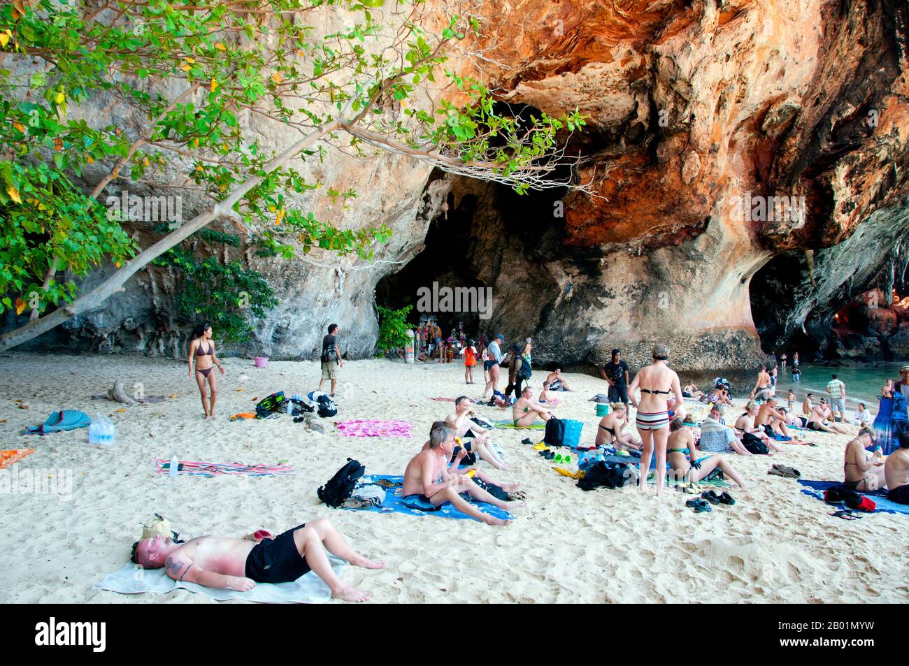 Thailand: Enjoying the shade of the cliff next to the Phra Nang cave, Tham Phra Nang, Krabi Coast.  Tham Phra Nang or the ‘Cave of the Revered Lady’ is located near the southern tip of the narrow peninsula that divides the beaches of Rai Leh East and Rai Leh West.  Dedicated to the memory of an eponymous Indian princess said to have been drowned offshore centuries ago, a fertility cult has developed around the cave and a small shrine within. The cave itself is packed with red-tipped phalluses placed here by fishermen in the hope of good catches. Stock Photo