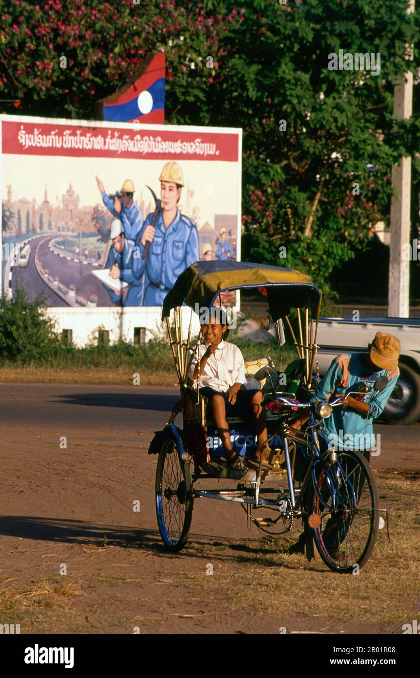 Laos: A pedicab or samlo in front of a revolutionary Socialist realist-style political poster on the streets of Vientiane.  Socialist realism is a style of realistic art which was developed in the Soviet Union and became a dominant style in other communist countries. Socialist realism is a teleologically-oriented style having its purpose the furtherance of the goals of socialism and communism. Although related, it should not be confused with social realism, a type of art that realistically depicts subjects of social concern. Stock Photo