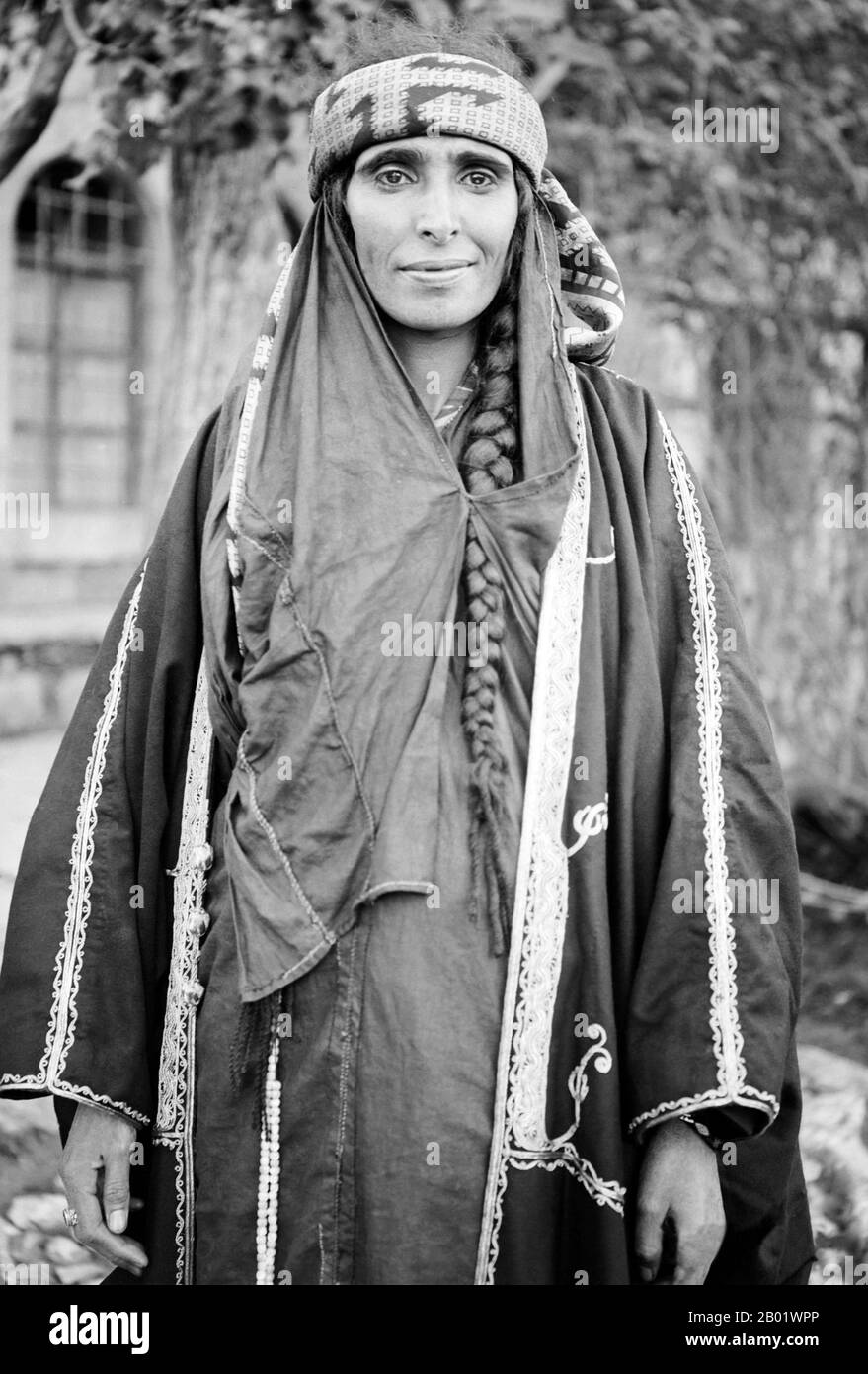 Jordan: A Bedouin woman wearing an embroidered gown, Wadi Musa, Petra, c. 1900s.  The Bedouin are a part of a predominantly desert-dwelling Arabian ethnic group traditionally divided into tribes or clans, known in Arabic as ʿašāʾir. The term 'Bedouin' means, 'those in bādiyah' or 'those in the desert'.   Starting in the late nineteenth century, many Bedouin under British rule began to transit to a semi-nomadic life. In the 1950s and 1960s, large numbers of Bedouin throughout Midwest Asia started to leave the traditional, nomadic life to settle in the cities. Stock Photo