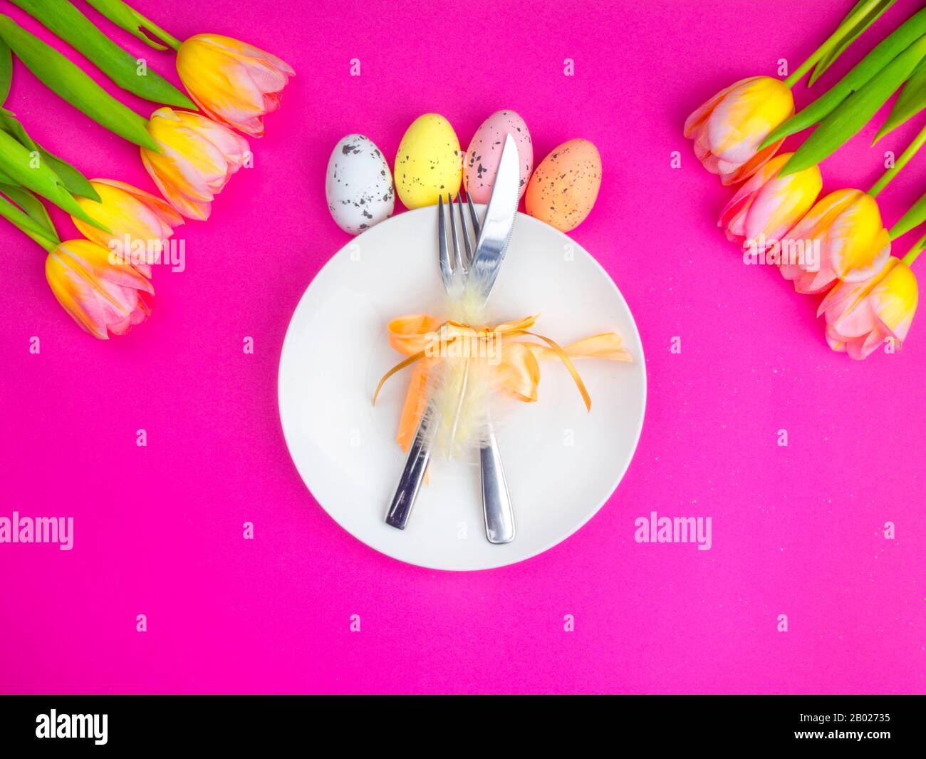 festive easter table cutlery with knife, fork, decorated with feather, bow, multi-colored eggs, yellow tulips flowers on pink, spring holiday background. Top view Stock Photo