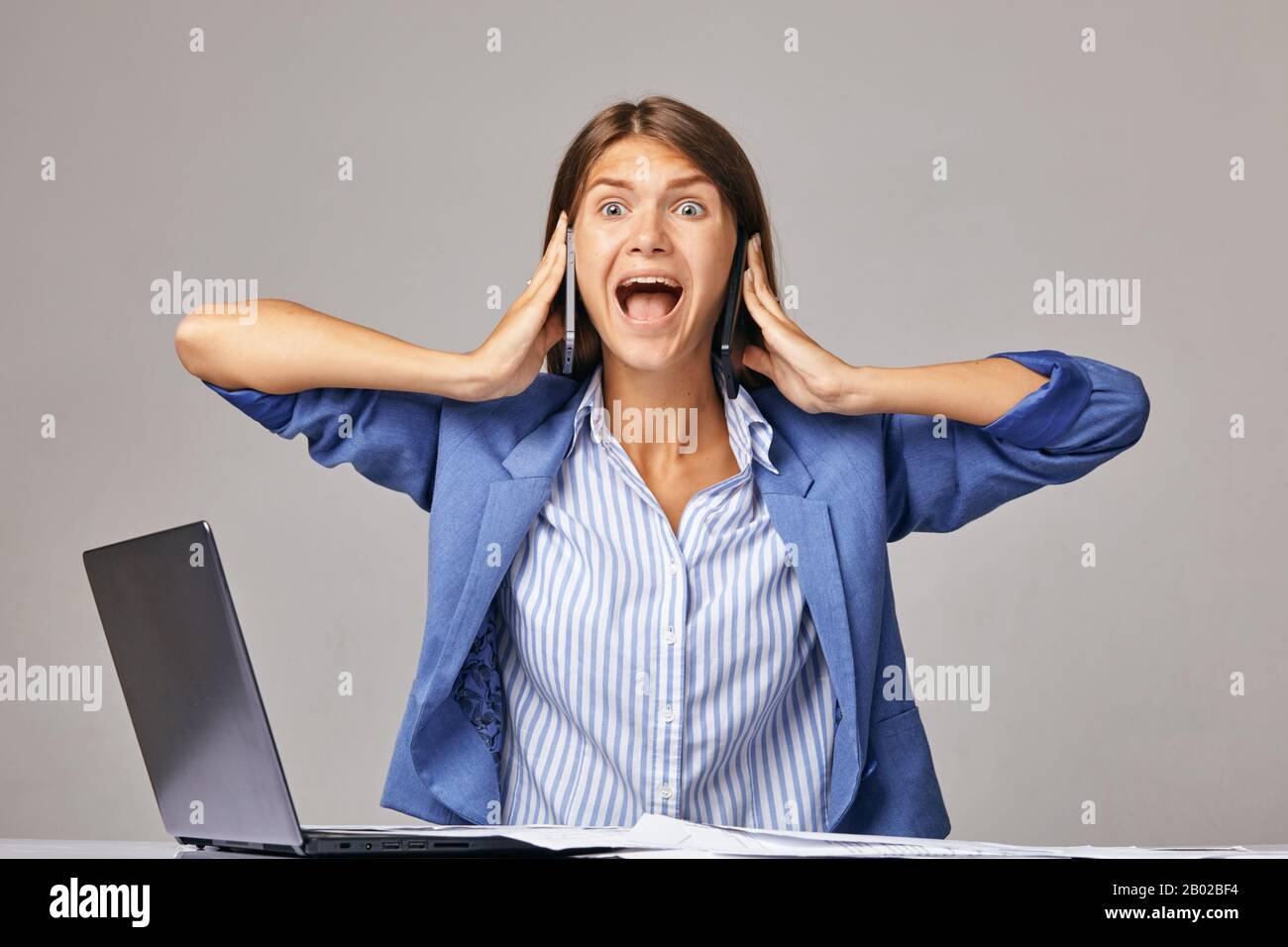 an angry businesswoman at workplace, talking on two smartphones at once Stock Photo