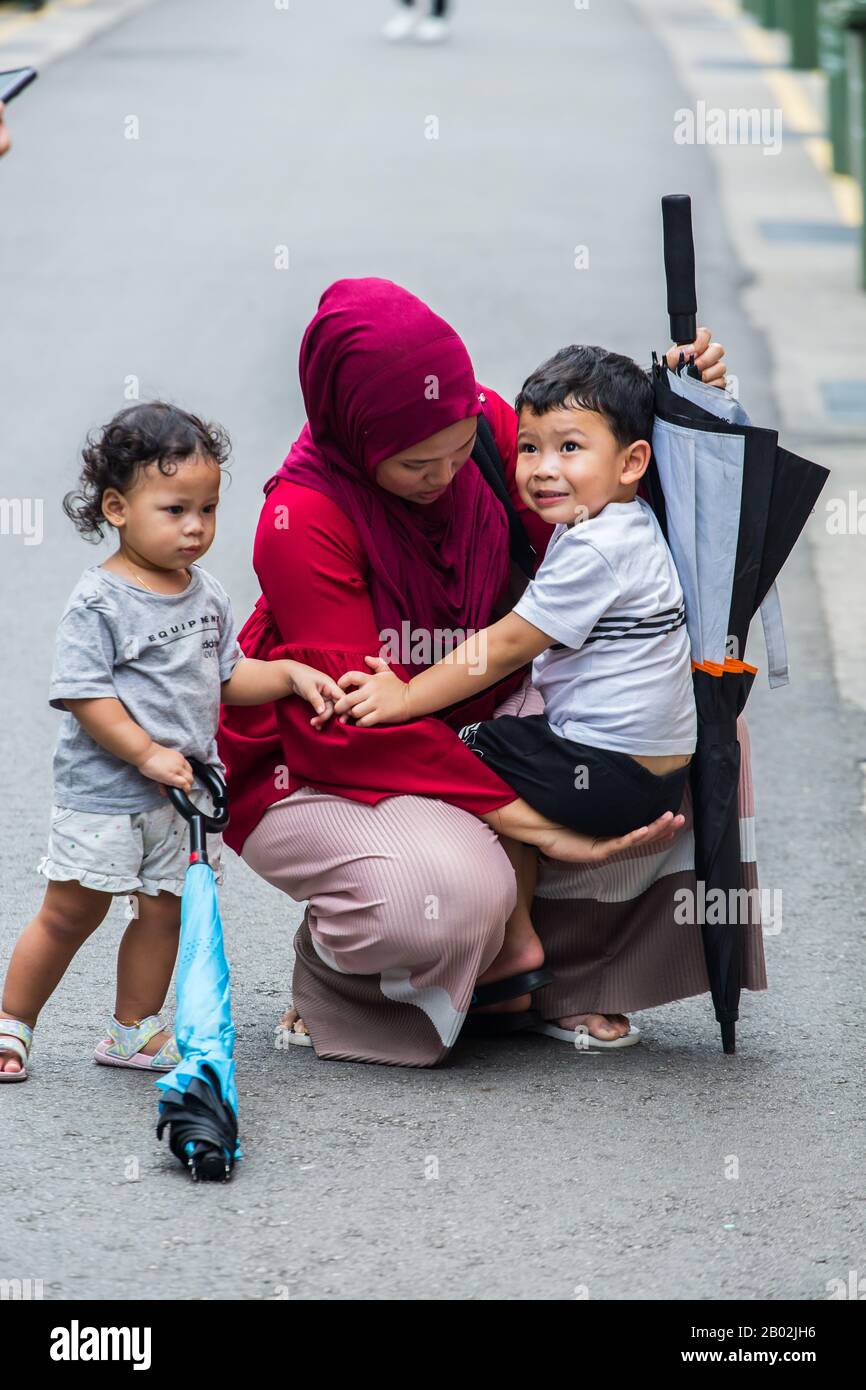 Vertical view of a busy Muslim mum in red hijab squat down to hold and support her crying child on the street. Singapore. Stock Photo