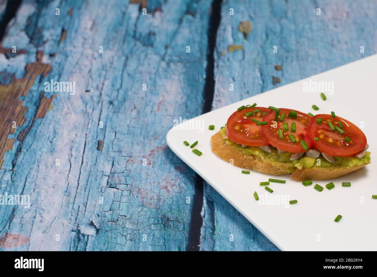 An open sandwich with avocado spread with garlic, sunflower kernels, slices of cherry tomatoes and diced chives, laid out on a white plate. Stock Photo