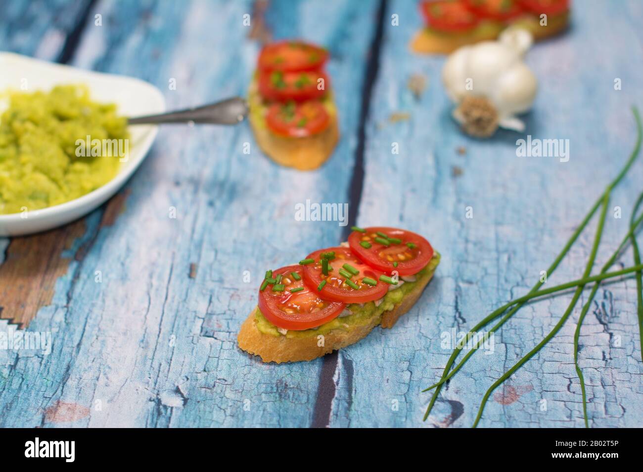 Open sandwich and a few chives scapes... two sandwiches, a bowl with avocado spread, and a garlic bulb are visible in a blurry background. Stock Photo