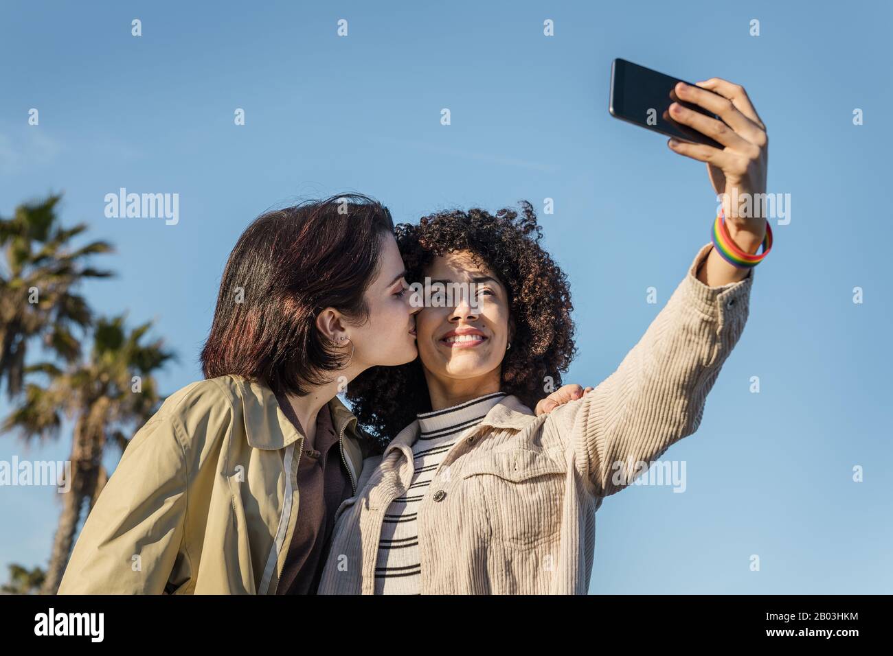 interracial young couple of homosexual women doing a selfie photo while kissing in a blue sky background with a mobile phone, concept of female friend Stock Photo