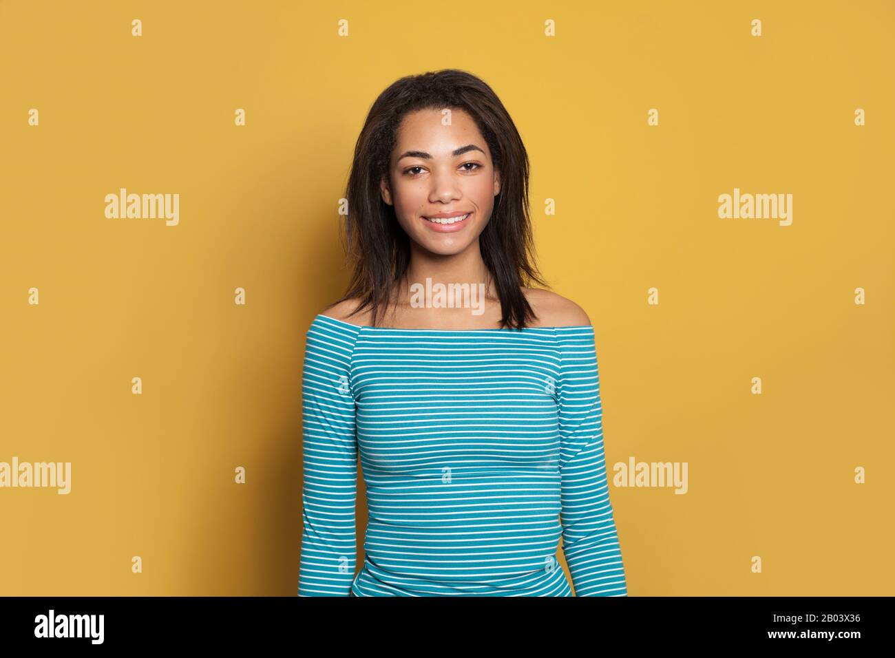 Happy smiling mixed race ethnicity black young woman in blue striped shirt on bright yellow studio background Stock Photo