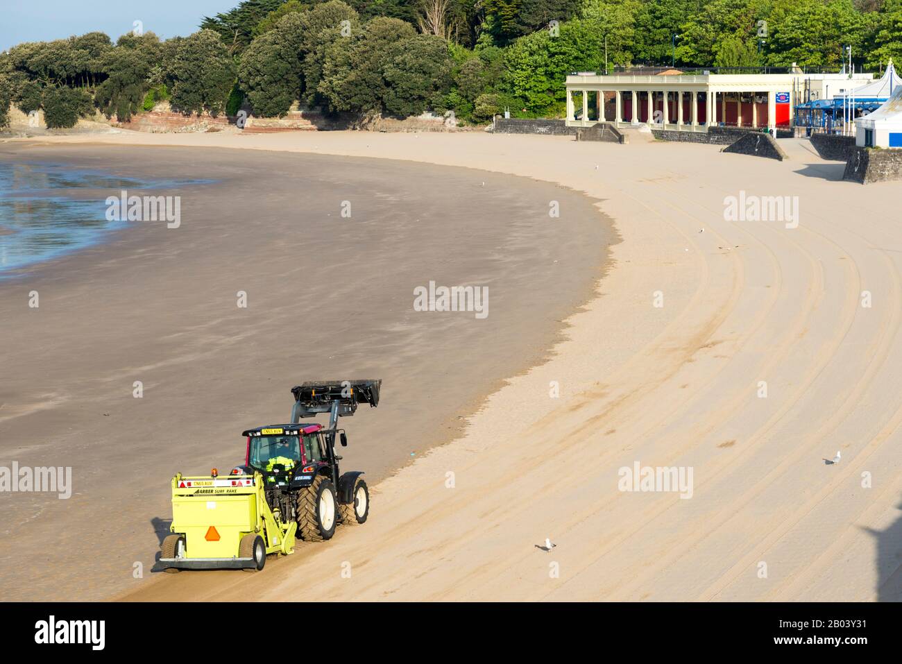Whitmore Bay beach at Barry Island, Wales,early on a bright sunny summer morning being cleaned of litter by a red Tractor pulling a Barber Surf Rake. Stock Photo