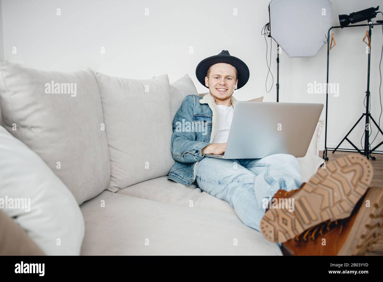 Freelancer photographer in hat video blogger works laptop, mounts videos for vlog about travel, in background lighting equipment Stock Photo