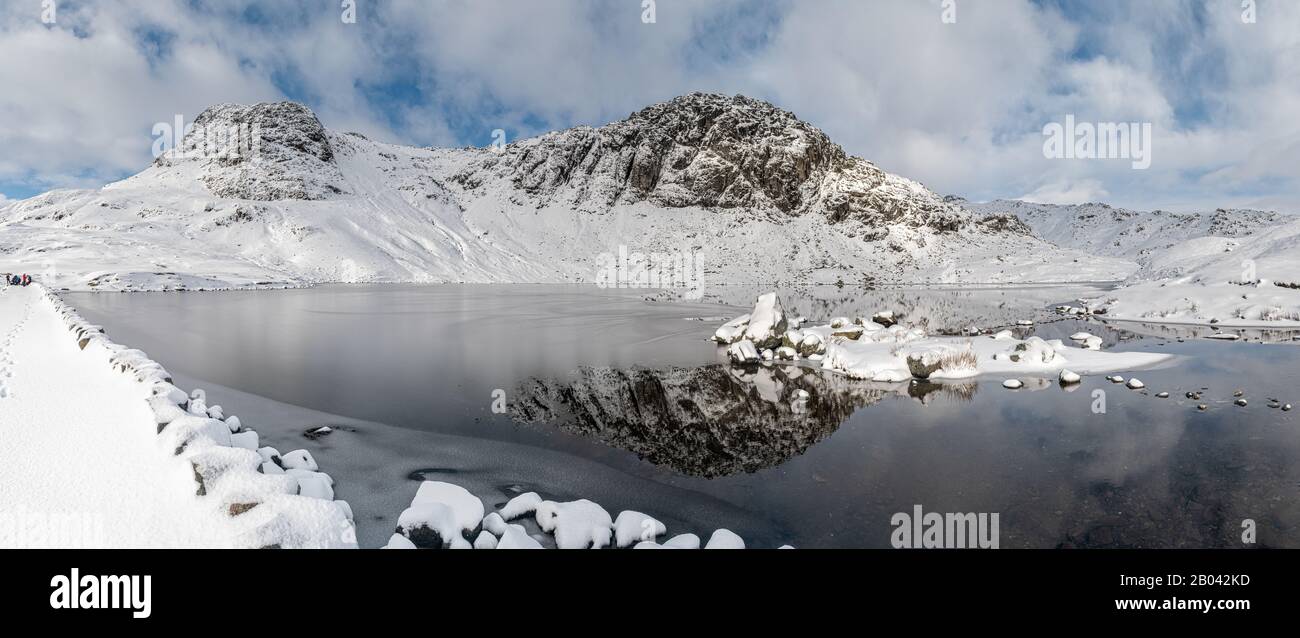 Panoramic view of Pavey Arc and Harrison Stickle reflected in a partially frozen Stickle Tarn Stock Photo