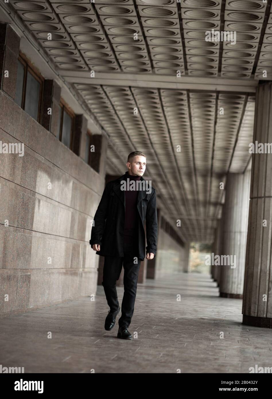 Stylish man in black trench coat and polo neck walks by the modernist building Stock Photo