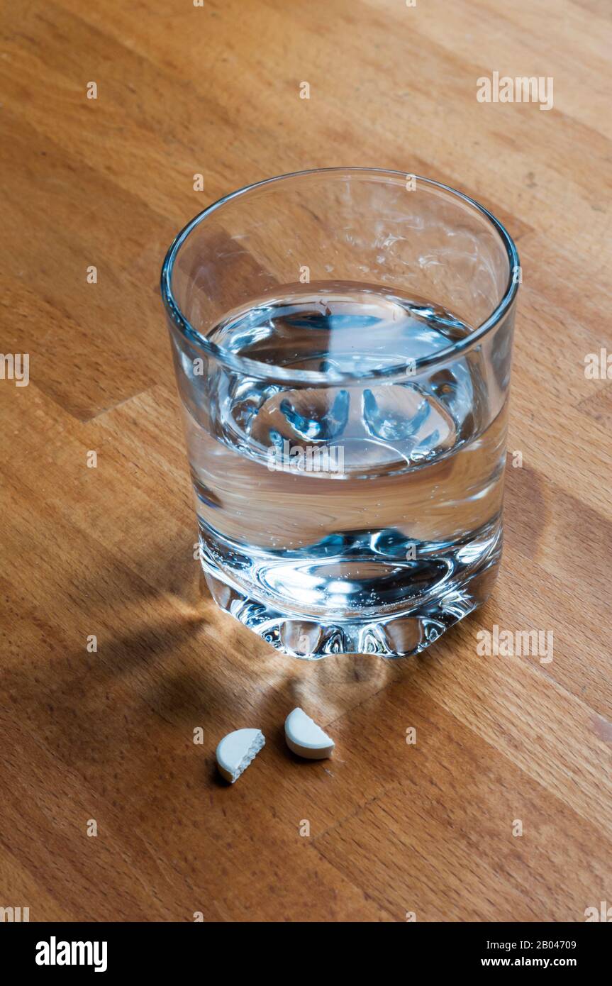 Painkiller tablet broken in half to administer half dosage amount, on bedside table with a glass of water. Stock Photo
