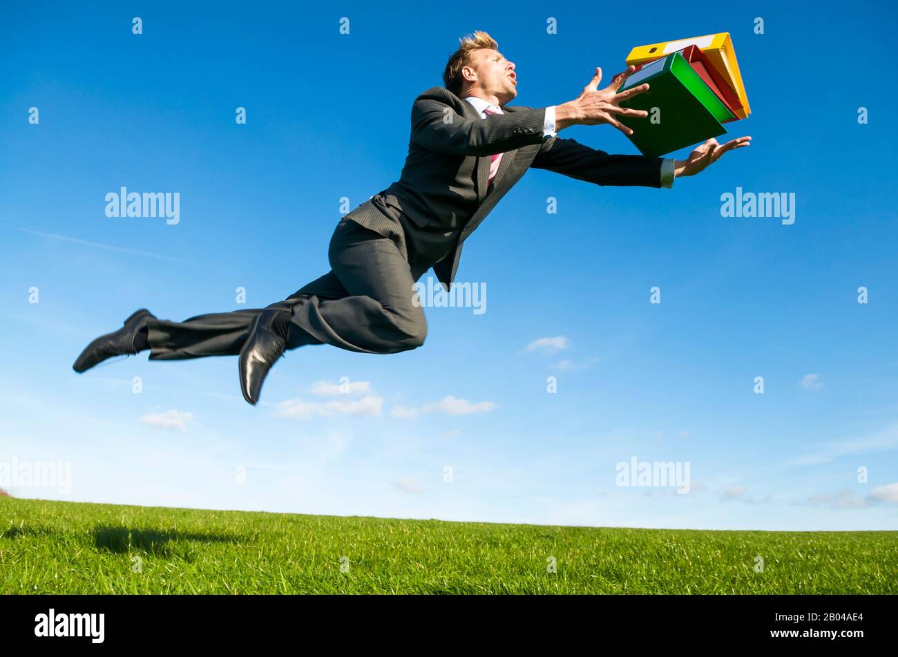 Careless office worker tripping dramatically with his file folders flying outdoors in a green grass field Stock Photo