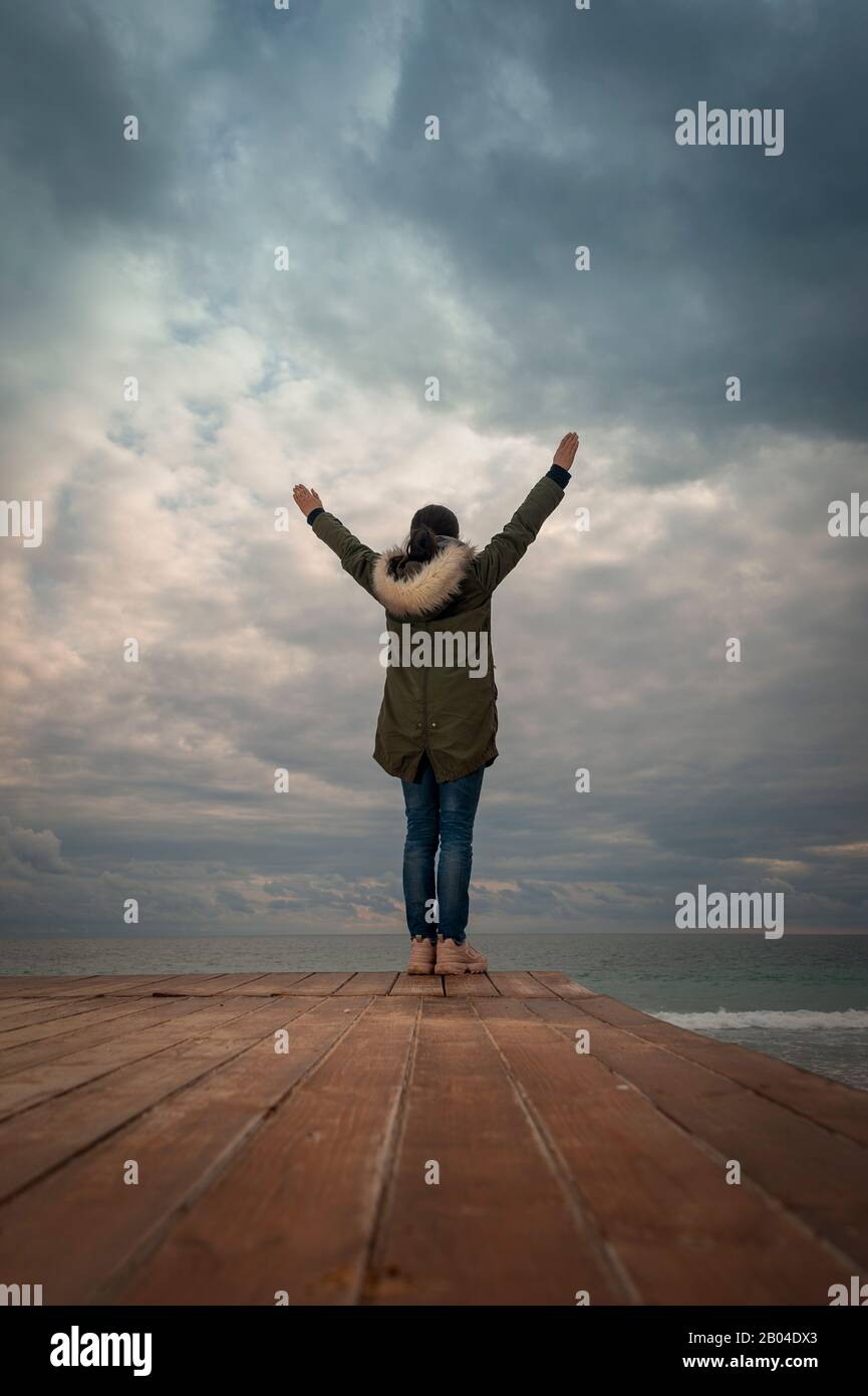 Woman wearing a Parka coat standing by the sea with her arms raised, lifestyle concept. Stock Photo