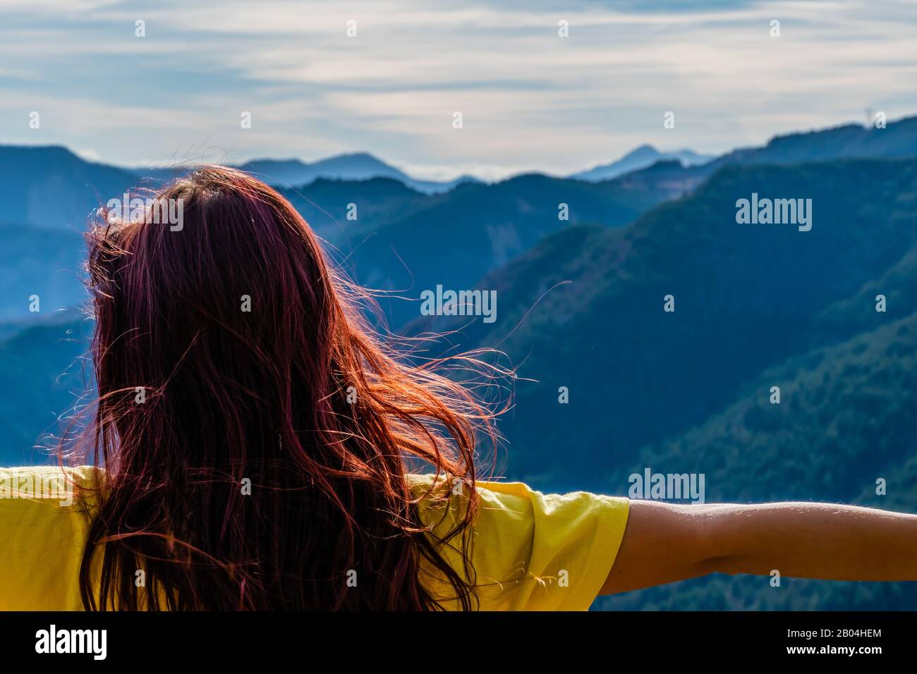 A close-up back view of a young redhead woman in front of the mountain landscape arms raised and hair fluttering from behind with no face on a sunny w Stock Photo
