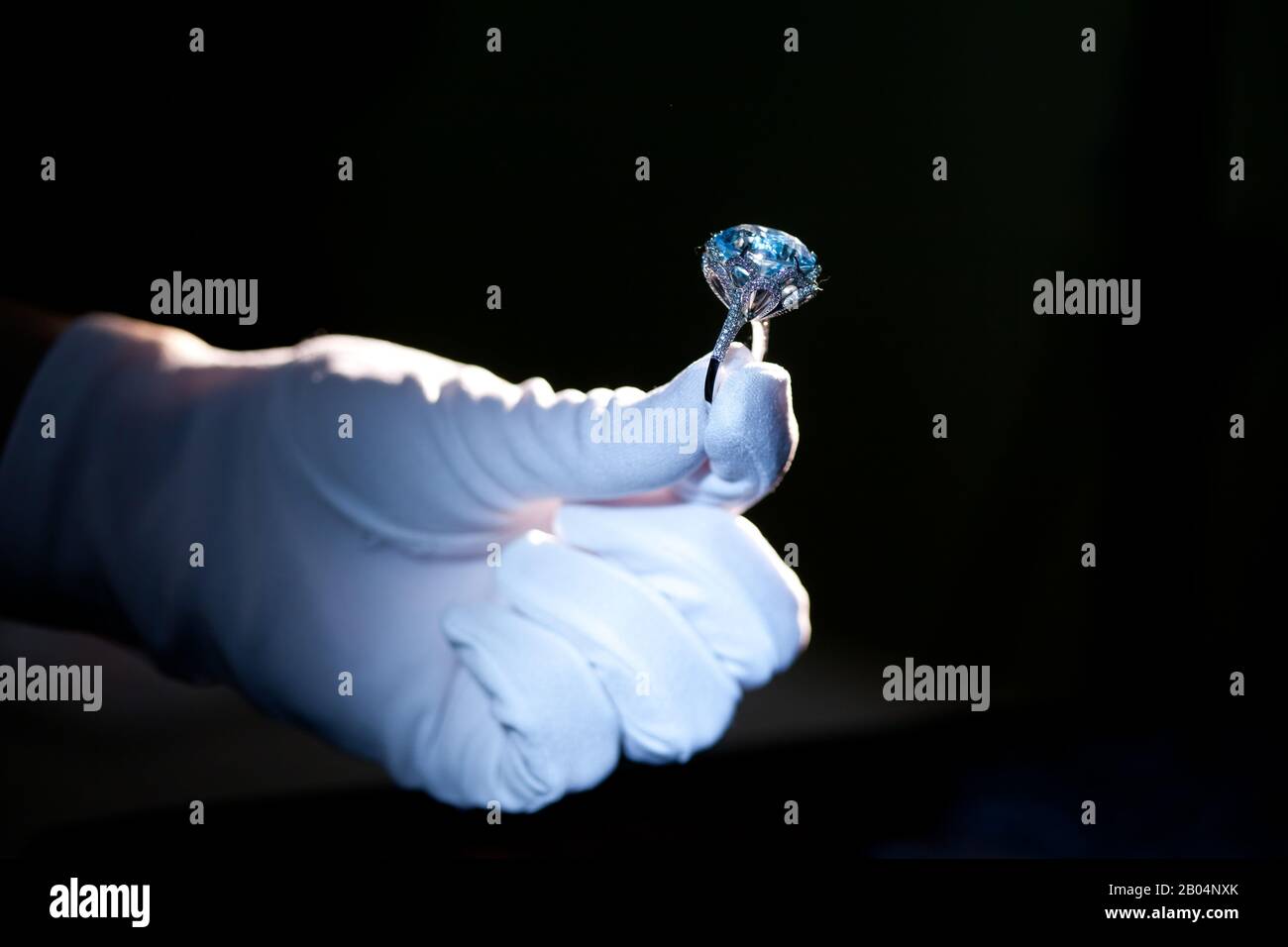 hand in white glove holding precious metal ring with big blue gem, on black background Stock Photo